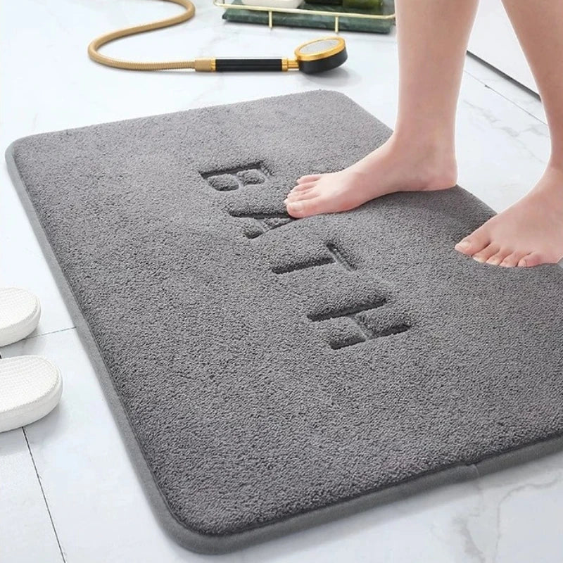 Plush grey bath mat with "BATH" embossed on its surface, a foot stepping onto it.